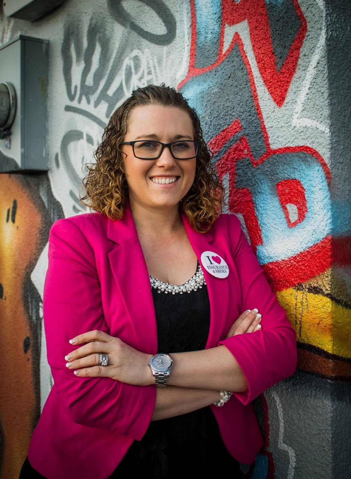 Woman in her thirties with curly hair wearing pink jacket and black dress with glasses