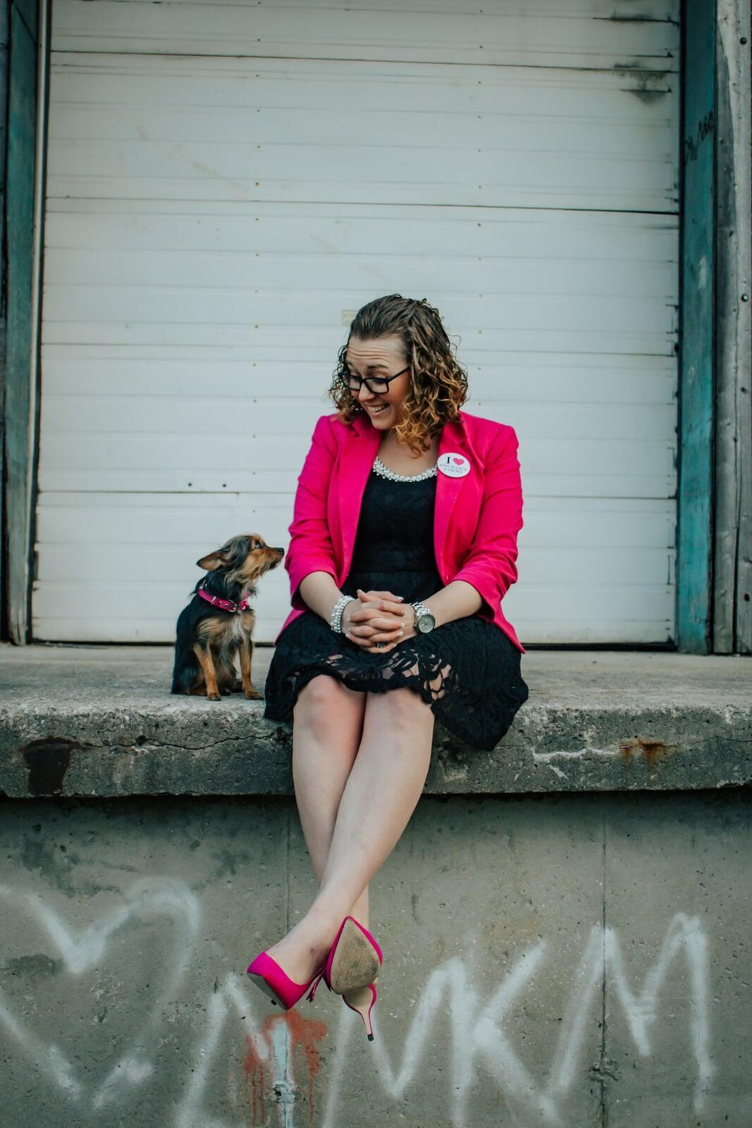 Woman with curly hair wearing glasses and pink jacket with black dress and small dog sitting beside her