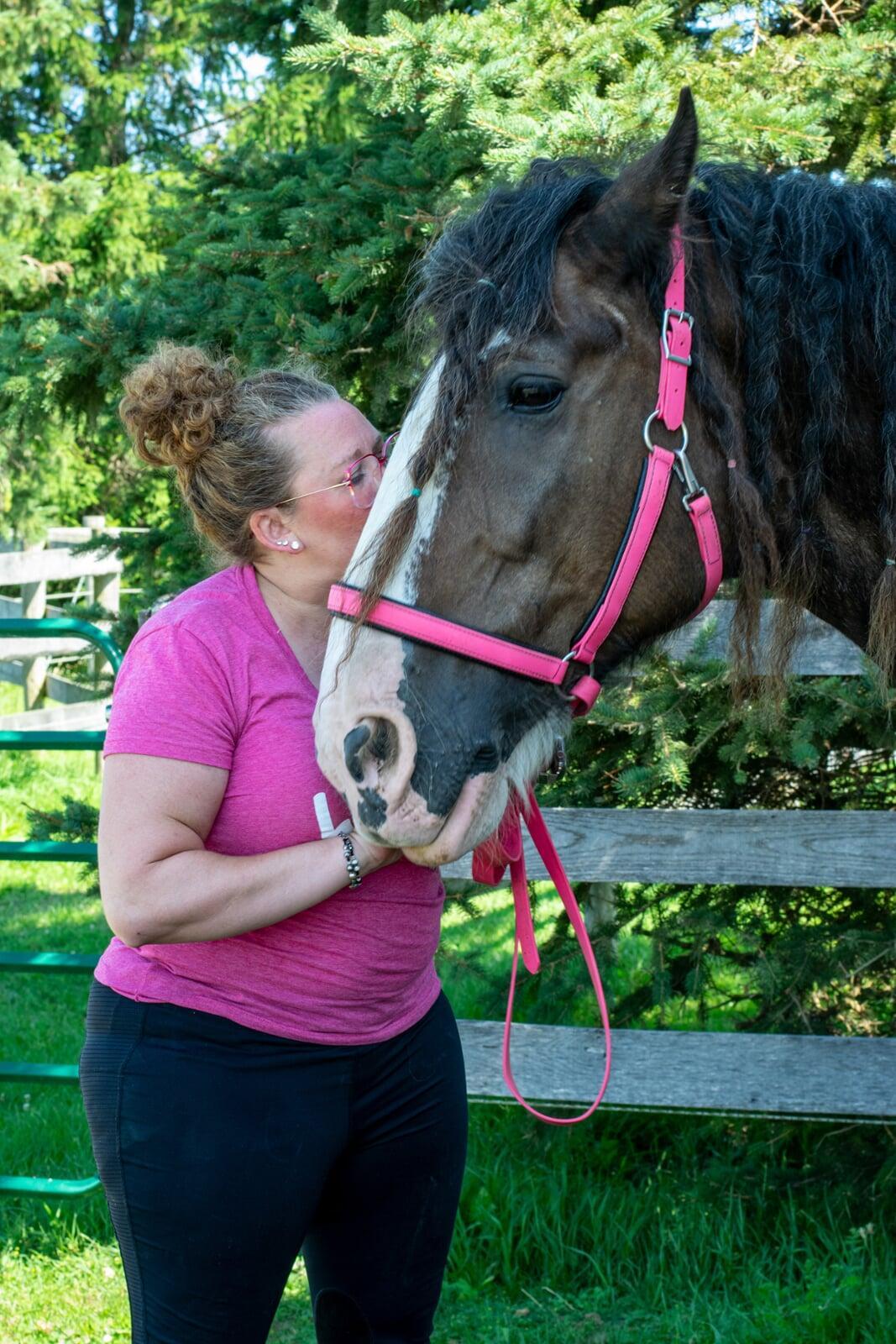 Woman in pink shirt and glasses kissing large horses head